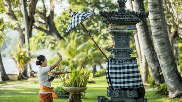 Uma pessoa realizando um ritual com um guarda-chuva xadrez e oferendas em uma estrutura de pedra tradicional em um jardim exuberante.