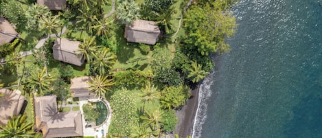 An aerial view of a coastal resort with thatched-roof cottages, palm trees, and a clear view of the ocean.
