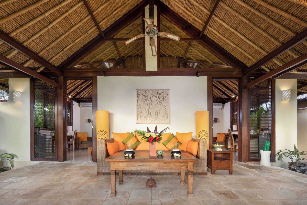 A covered outdoor seating area with a wooden table and chairs, a ceiling fan, and a decorative wall piece.