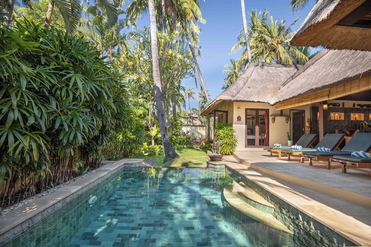 A pool area with lounge chairs, a thatched-roof building, and lush tropical vegetation.