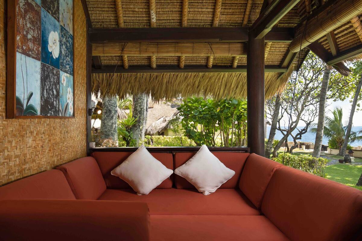 A red sofa with cushions, a woven wall, and a thatched roof structure.