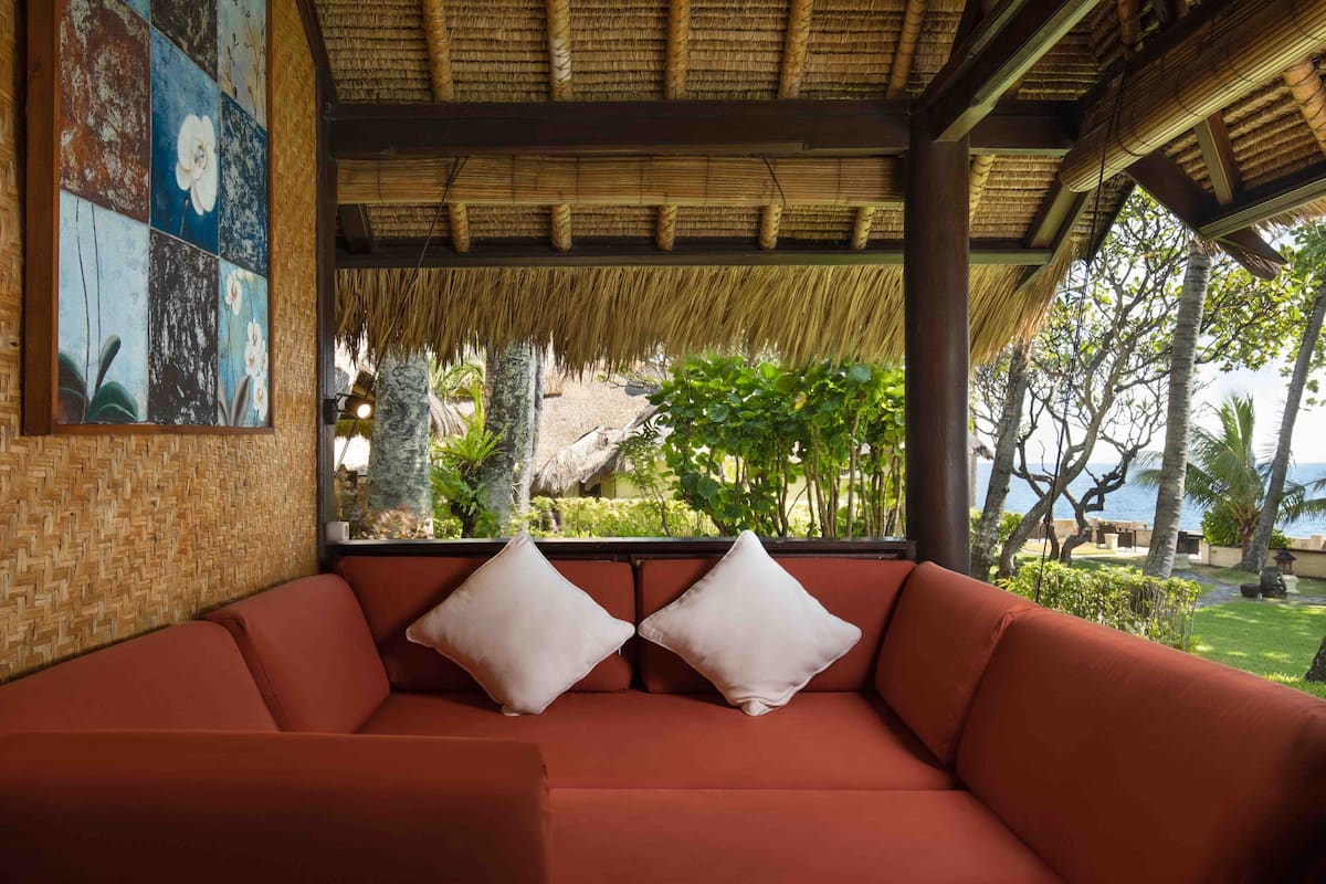 A red sofa with two pillows, a woven wall, and a thatched roof structure.