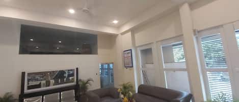 Un salón amplio con un televisor de pantalla plana grande, dos sofás grises, una mesa de centro y varias plantas en macetas.