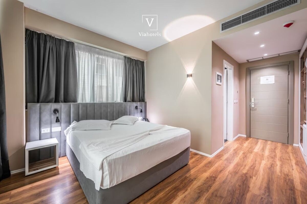 A modern hotel room with a large bed, a bedside table, a nightstand, a wall-mounted light, and a door.