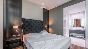 A modern bedroom with a tufted headboard, bedside lamps, a large bed, and a bathroom with a vanity and mirror. - Vh Eurostar Tirana Hotel Congress & Spa (Tirana)