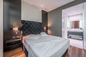 A modern bedroom with a tufted headboard, bedside lamps, a large bed, and a bathroom with a vanity and mirror. - Vh Eurostar Tirana Hotel Congress & Spa (Tirana)
