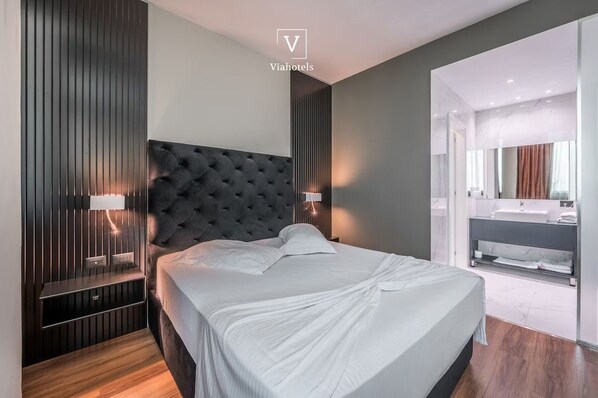 A modern bedroom with a tufted headboard, bedside lamps, a large bed, and a bathroom with a vanity and mirror. - Vh Eurostar Tirana Hotel Congress & Spa (Tirana)