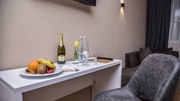 Ein Hotelzimmer mit einer weißen Minibar, einem an der Wand befestigten Flachbildfernseher, einem grau gepolsterten Sessel und einem Fenster mit dunklen Vorhängen.