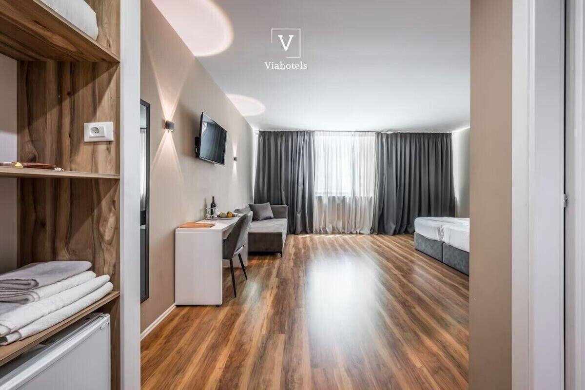 A modern hotel room with a wooden floor, a desk, a sofa, a bed, and a TV mounted on the wall.