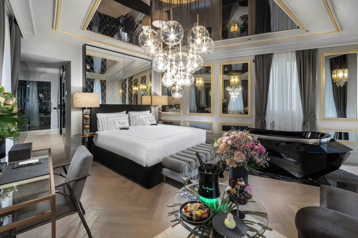 A luxurious hotel room with a large bed, a piano, a dining table with a cake and flowers, and a chandelier.