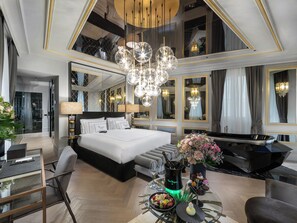Ein luxuriöses Hotelzimmer mit einem großen Bett, einem Klavier, einem Esstisch mit Kuchen und Blumen sowie einem Kronleuchter.