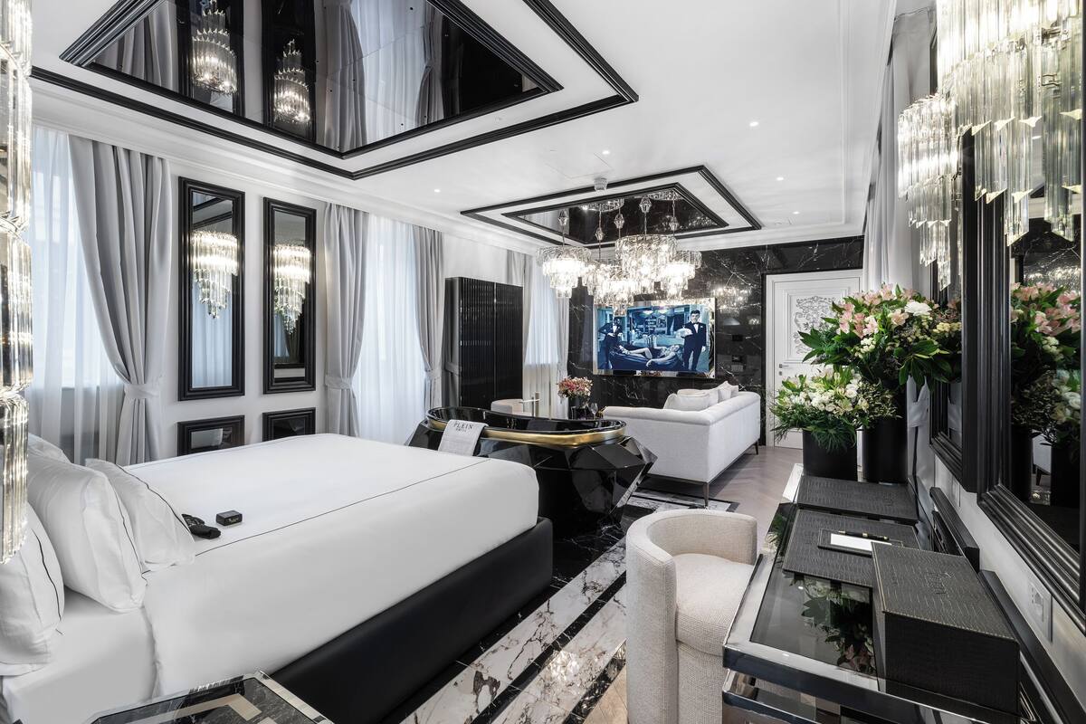 A modern hotel room with a large bed, a sitting area with a sofa and a small table, a TV, and a mirror with floral decorations.