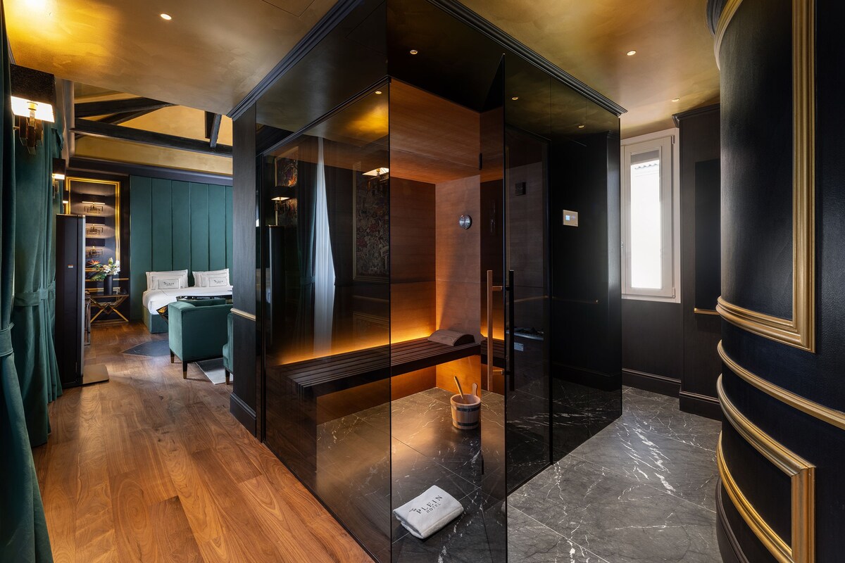 A modern hotel room with a glass-enclosed seating area, wooden flooring, and a view of the cityscape.
