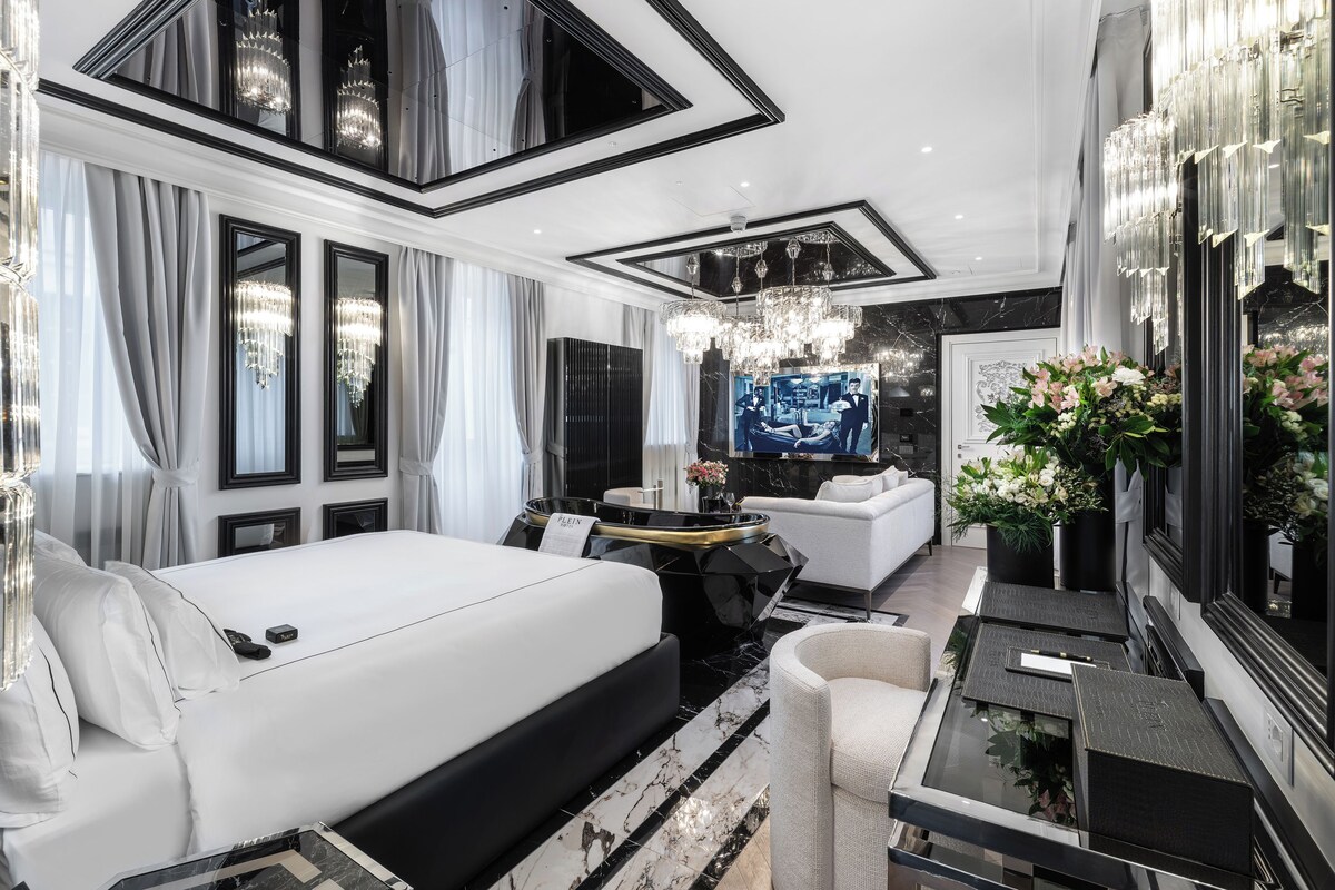 A modern hotel room with a large bed, a sitting area with a sofa and a small table, a TV, and a mirror with floral decorations.