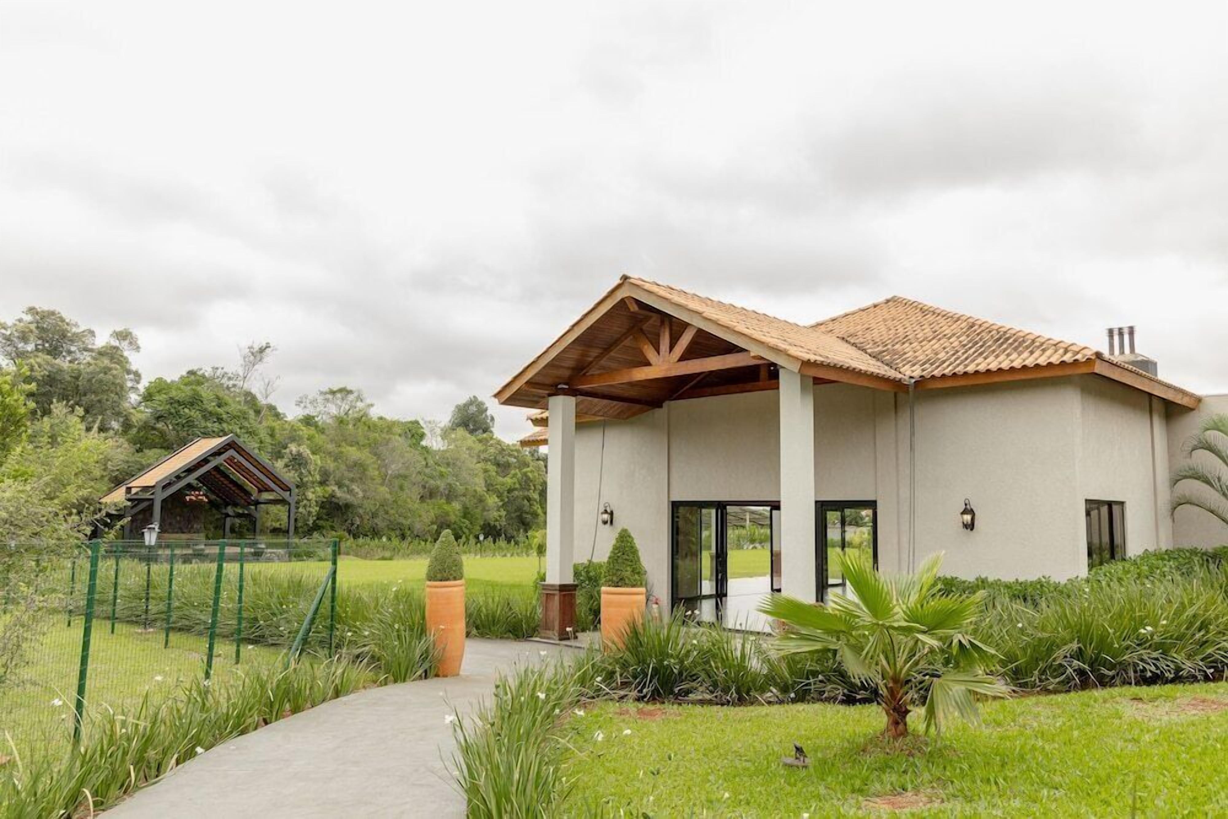 Um edifício moderno com entrada coberta, jardim cercado e uma pequena construção ao fundo.