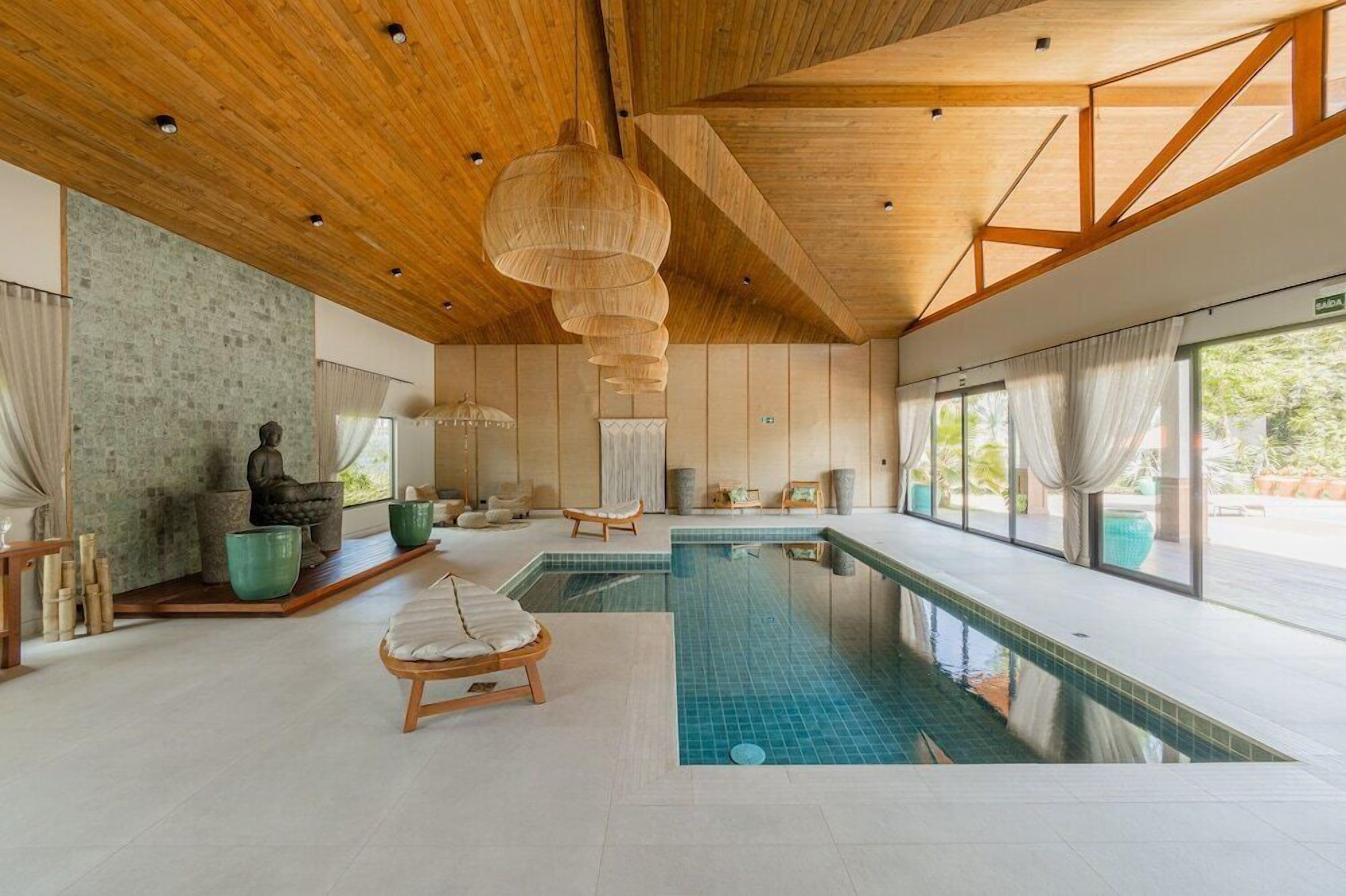 Piscina interior moderna com vigas de madeira, uma escultura de pedra e vista para o verde lá fora.