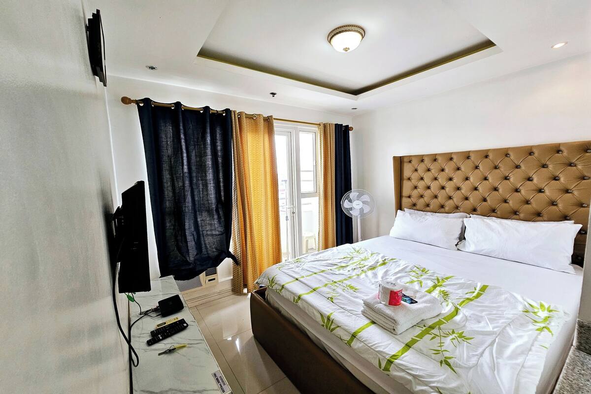 Ein Hotelzimmer mit einem großen Bett, einem gepolsterten Kopfteil, einem Fenster mit Vorhängen und einem Deckenventilator.