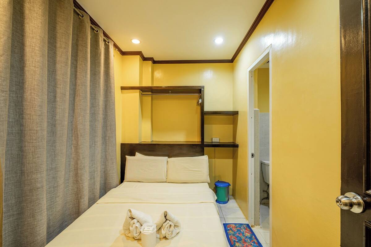 A hotel room with a bed, a small table, a blue trash bin, and a door.