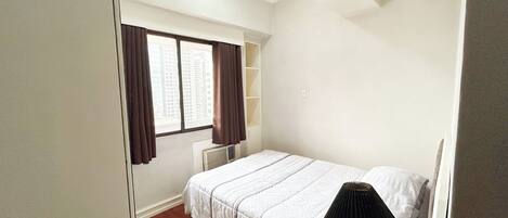 A bedroom with a bed, a nightstand, a window with curtains, and a radiator.
