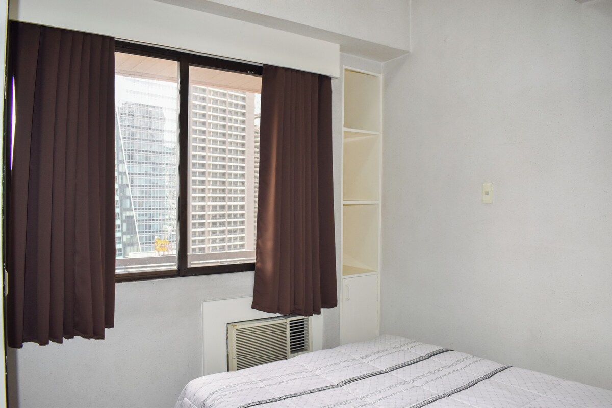 A bedroom with a bed, a window with curtains, a radiator, and a view of a building outside.
