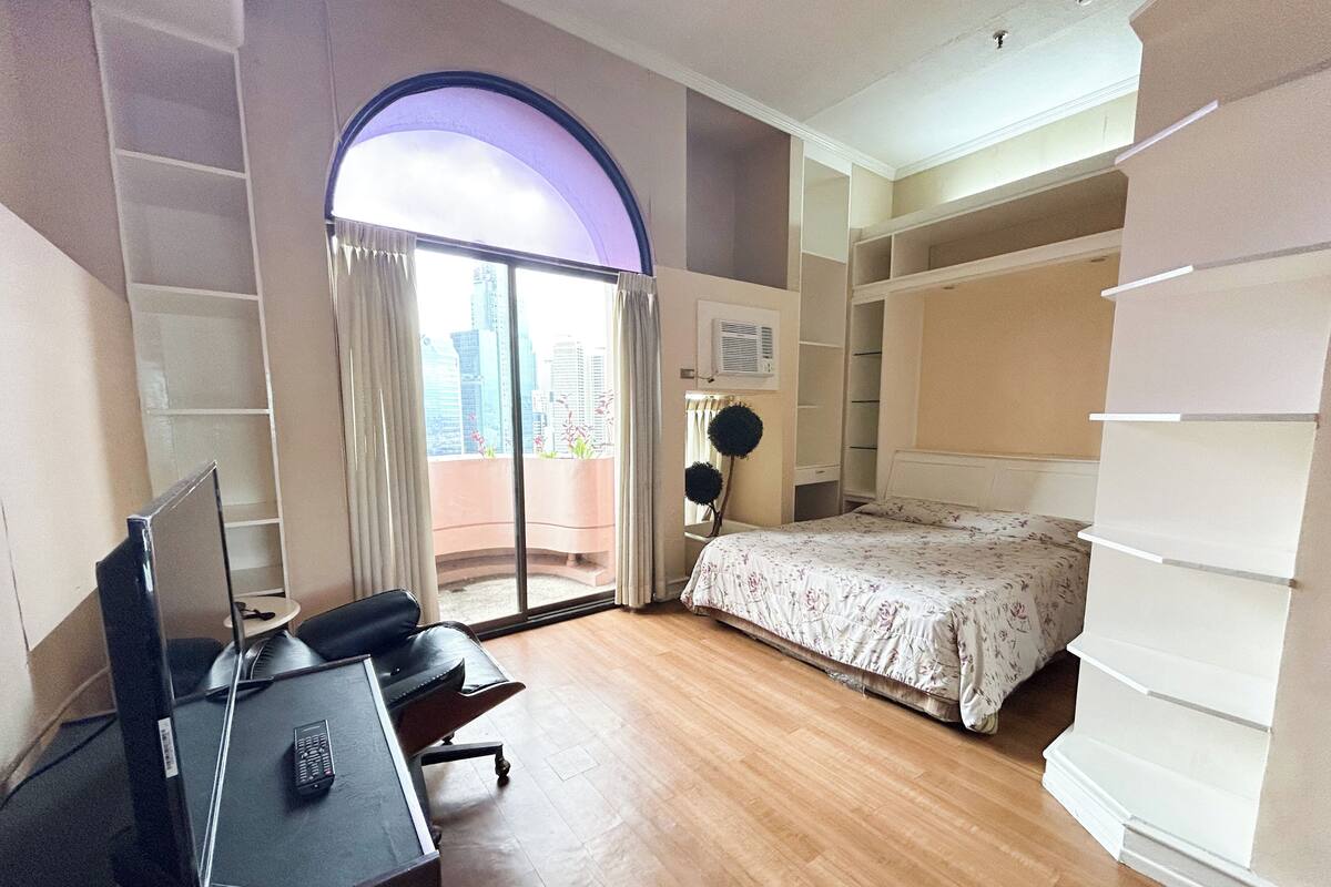 A bedroom with a bed, a desk with a computer, a chair, a wardrobe, and a view of the city through a large window.