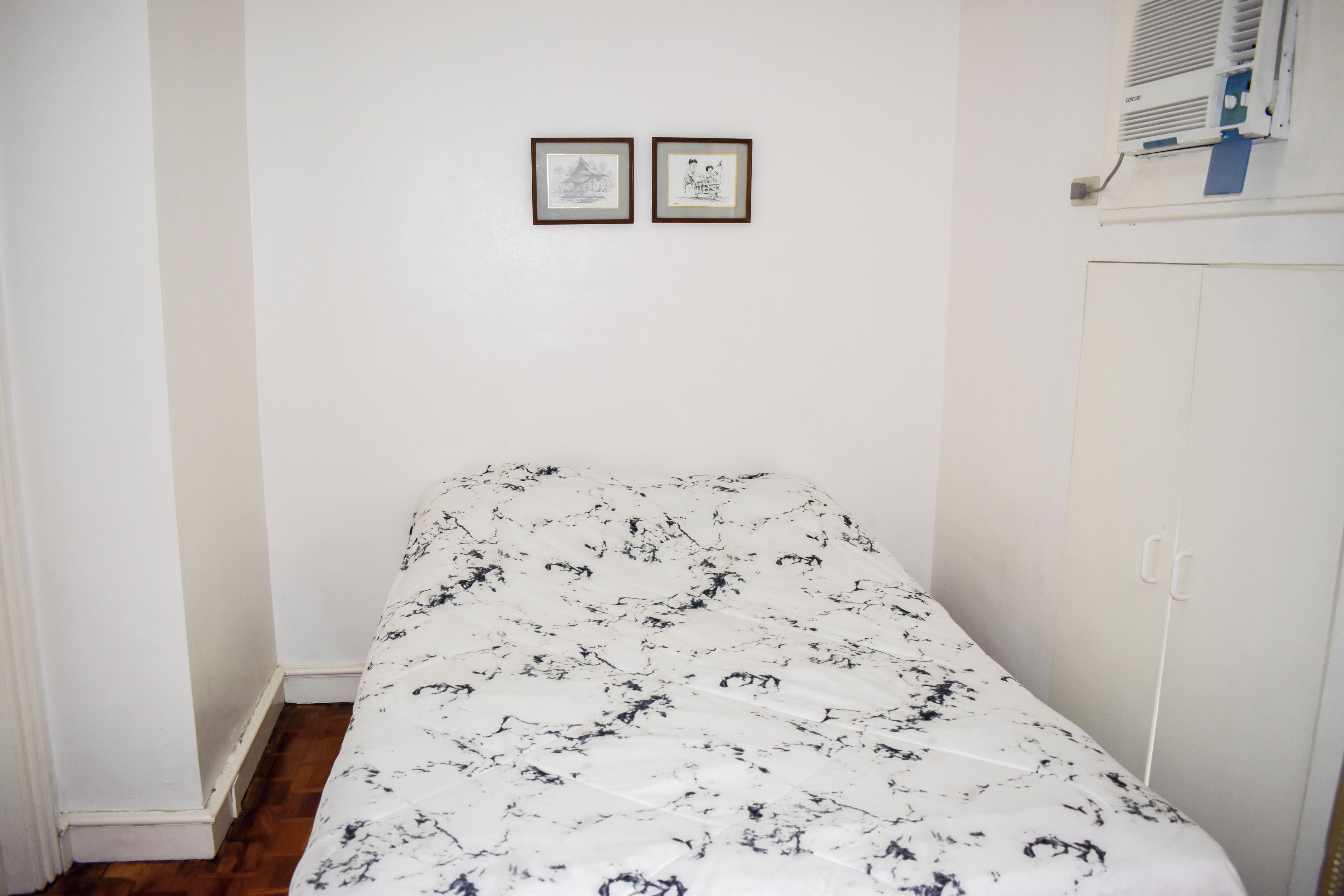 Een kleine kamer met een enkel bed, een witte muur en een houten vloer. Twee ingelijste foto’s hangen aan de muur, er is een witte garderobe en een witte airconditioning unit boven de garderobe.