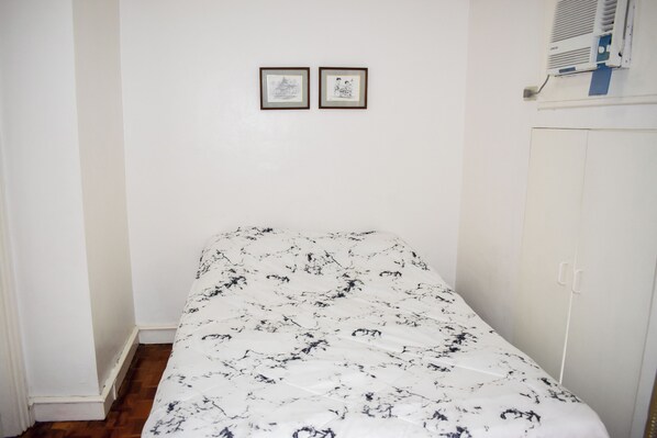 Quarto pequeno com cama de solteiro, parede branca e chão de madeira. Há duas imagens emolduradas na parede, um guarda-roupa branco e um aparelho de ar condicionado branco sobre o guarda-roupa.