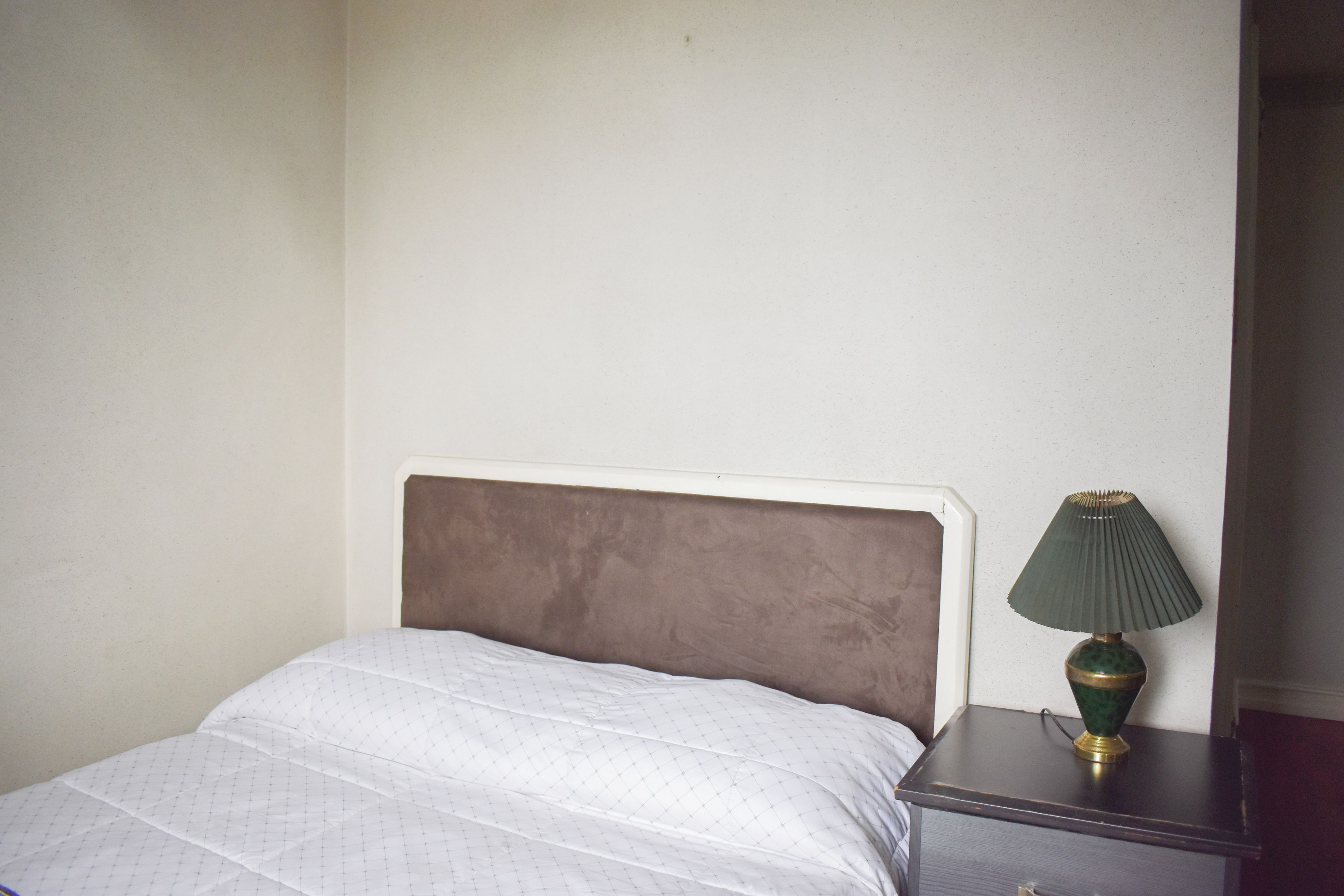 Un lit avec une couette blanche et une tête de lit marron, une table de chevet avec une lampe verte et un mur sobre.