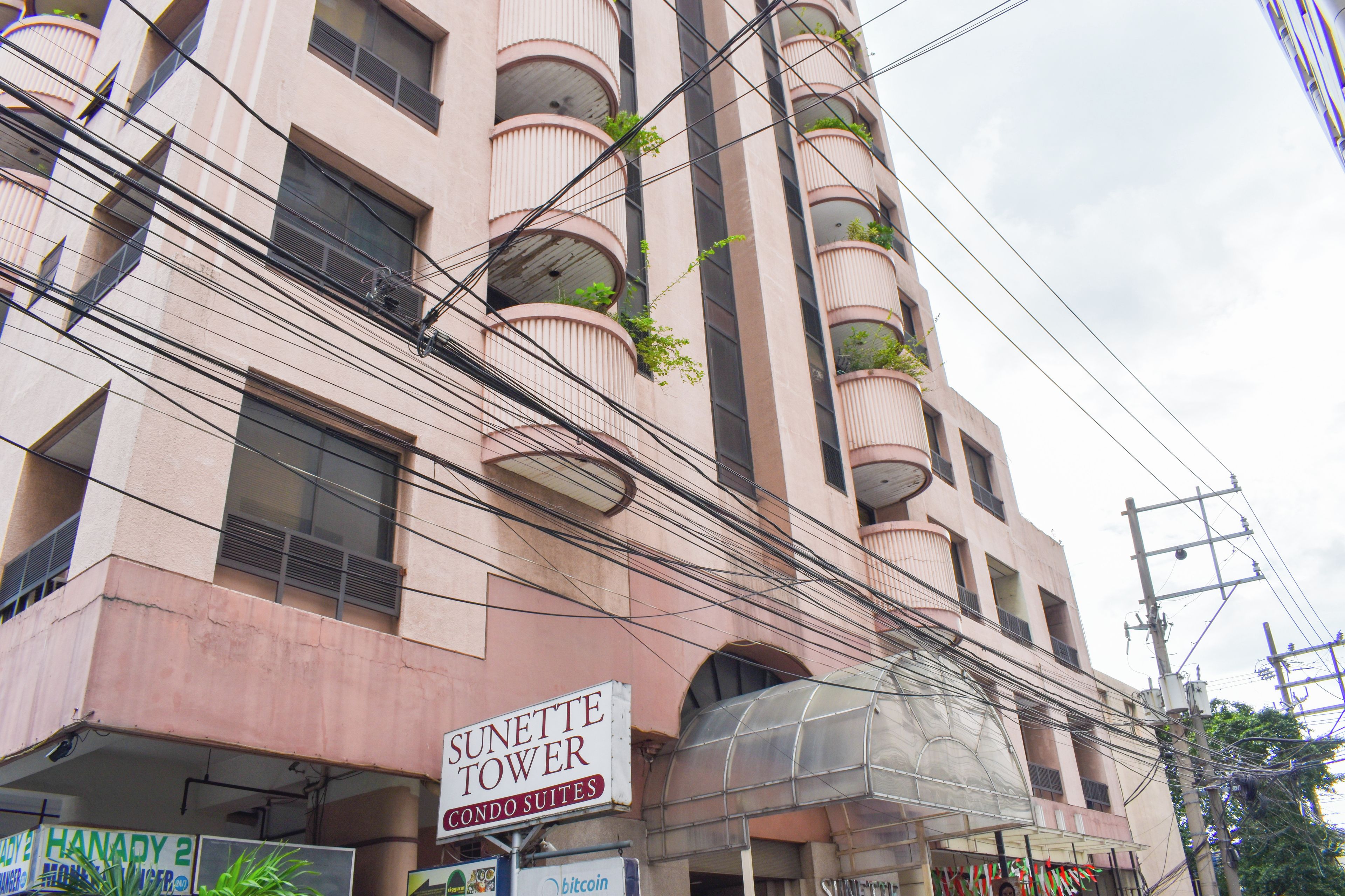 Un immeuble à plusieurs étages doté de balcons, une enseigne indiquant « Sunette Tower Condo Suites », et divers autres panneaux, dont un portant l’inscription « HANADY 2 ».
