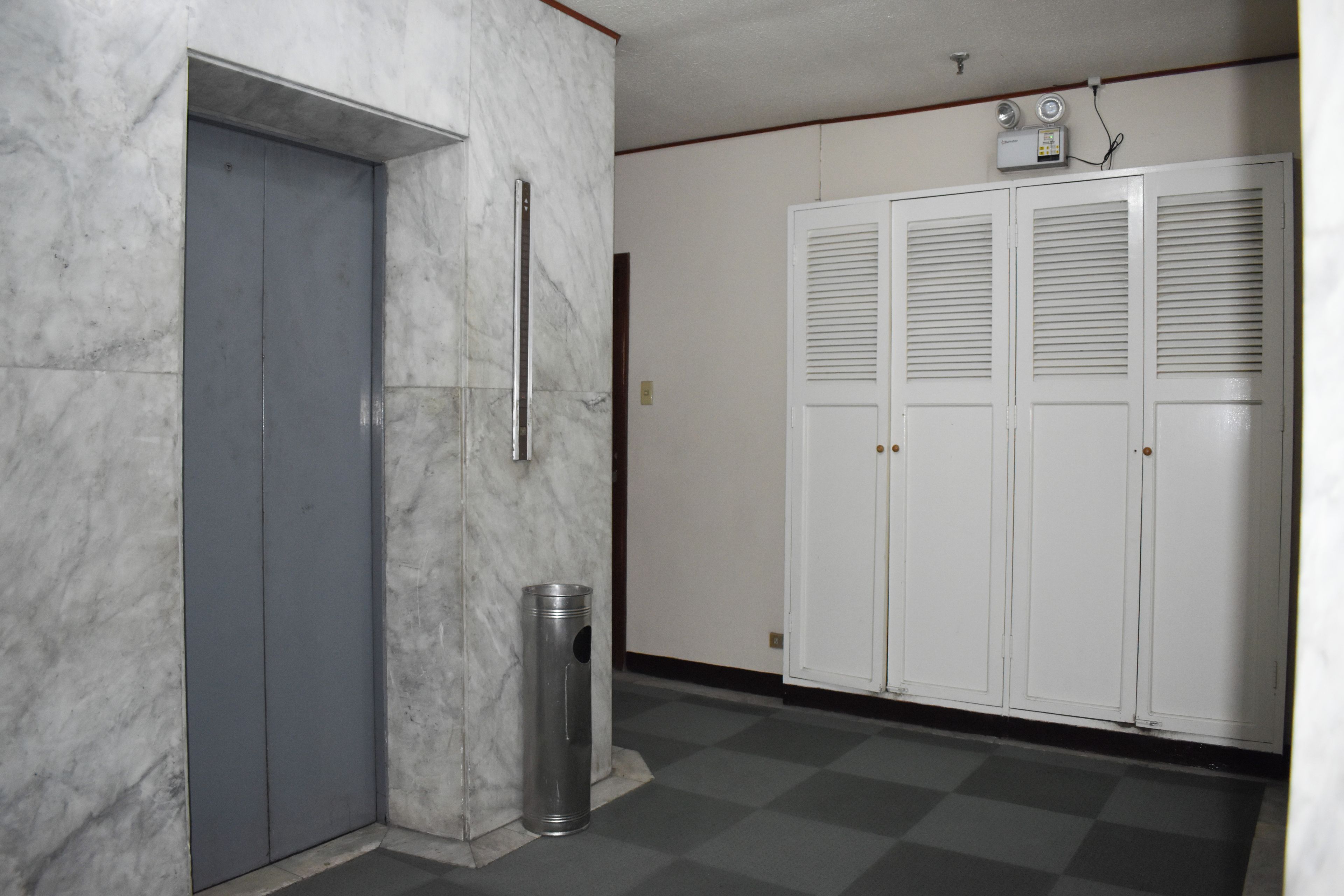 Un couloir avec un mur de marbre, un ascenseur gris, une poubelle et une rangée de casiers blancs.