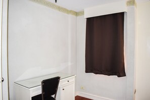 Um quarto com uma escrivaninha de vidro, uma cadeira preta, uma parede branca e uma cortina marrom.
