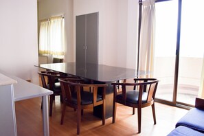 Een eetgedeelte met een glazen tafel, omringd door houten stoelen, een witte tafel aan de linkerkant en een grote kast op de achtergrond.