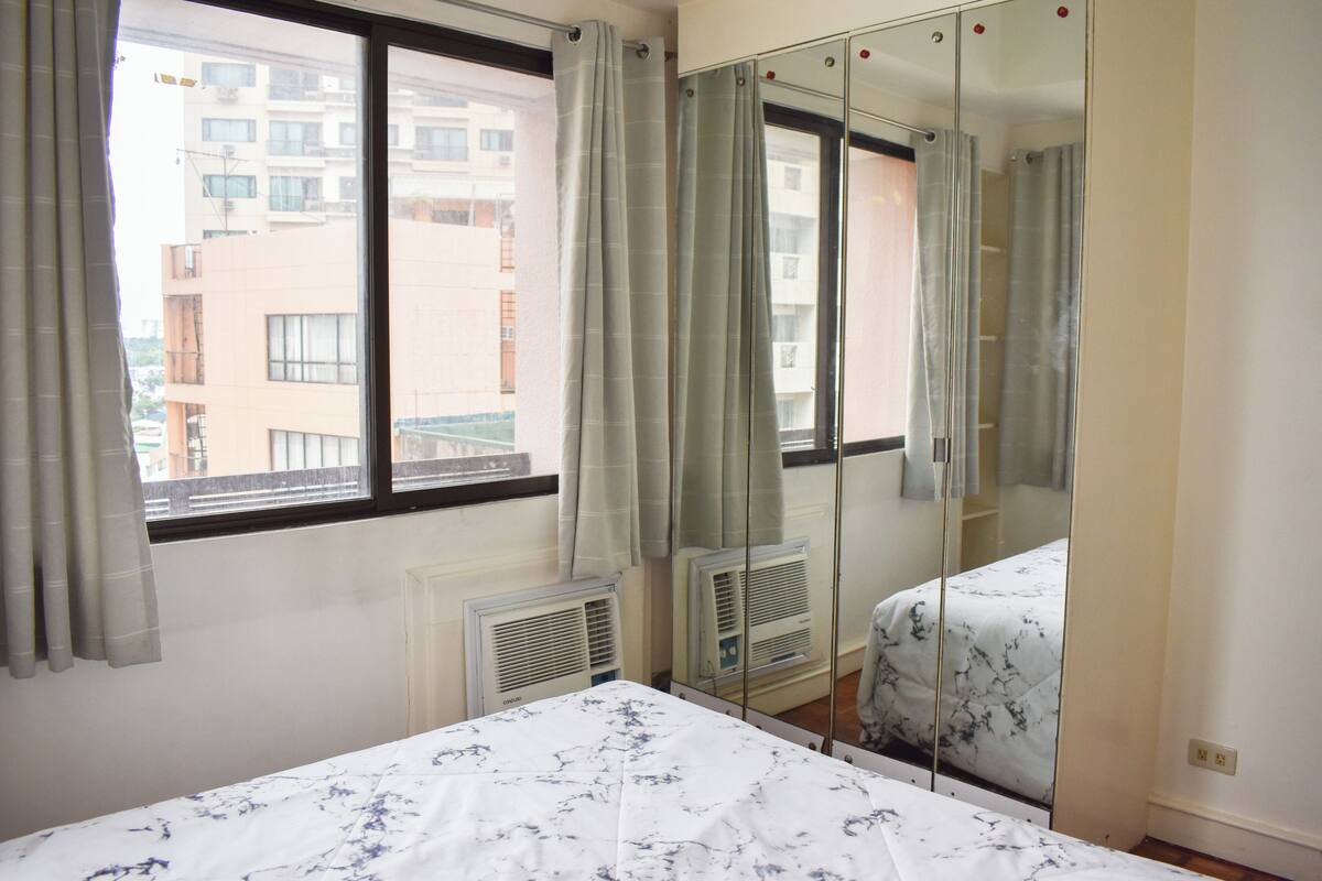 A bedroom with a bed, a window with curtains, a radiator, and a mirror.