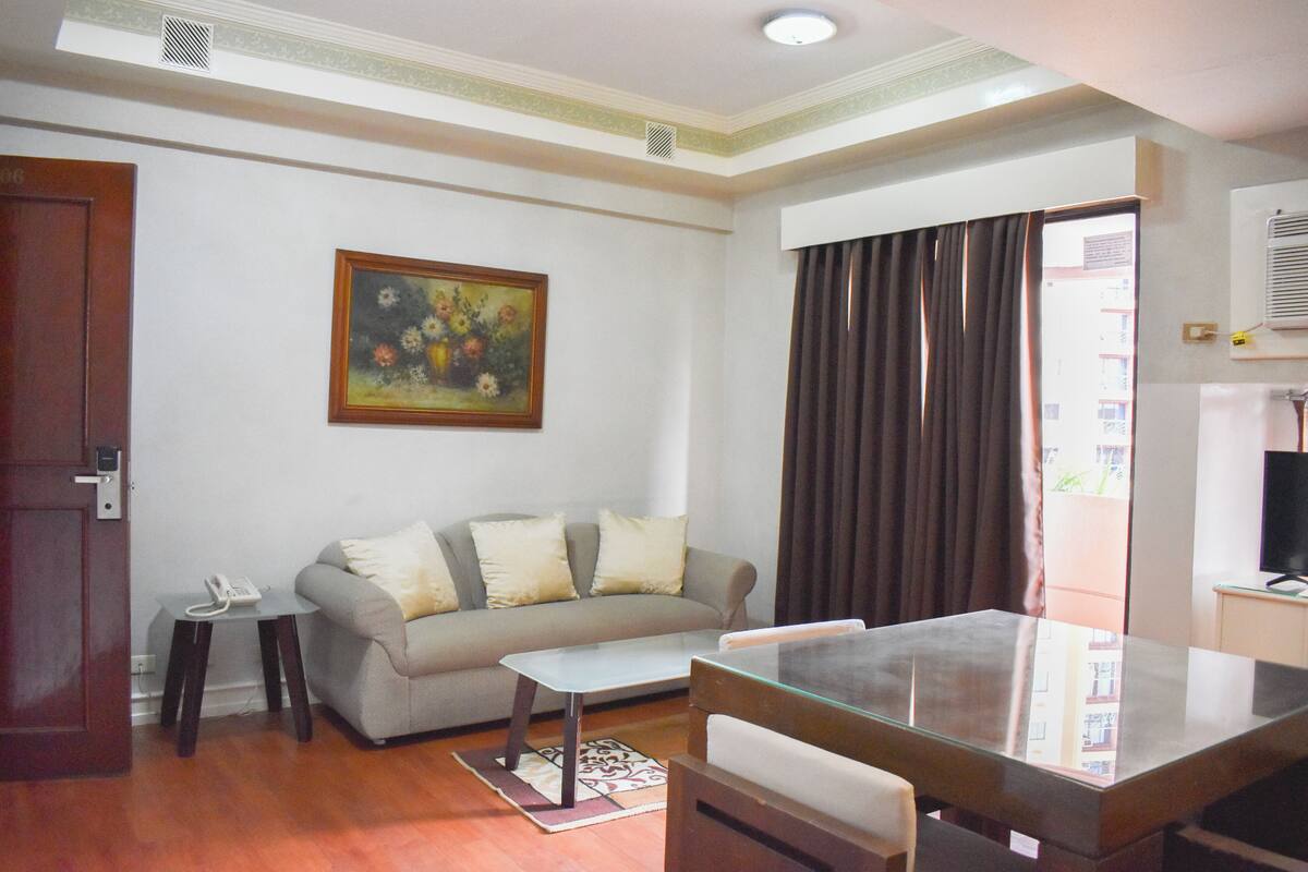A living room with a sofa, a coffee table, a dining table, a painting, a telephone, and a window with curtains.