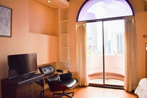 Habitación de hotel con televisión de pantalla plana, sillón de cuero, una mesita pequeña y un amplio ventanal corredero que da a un balcón.