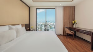 A hotel room with a large bed, a desk, and a view of a cityscape through a large window. - Palazzo 3 (Da Nang Municipality)