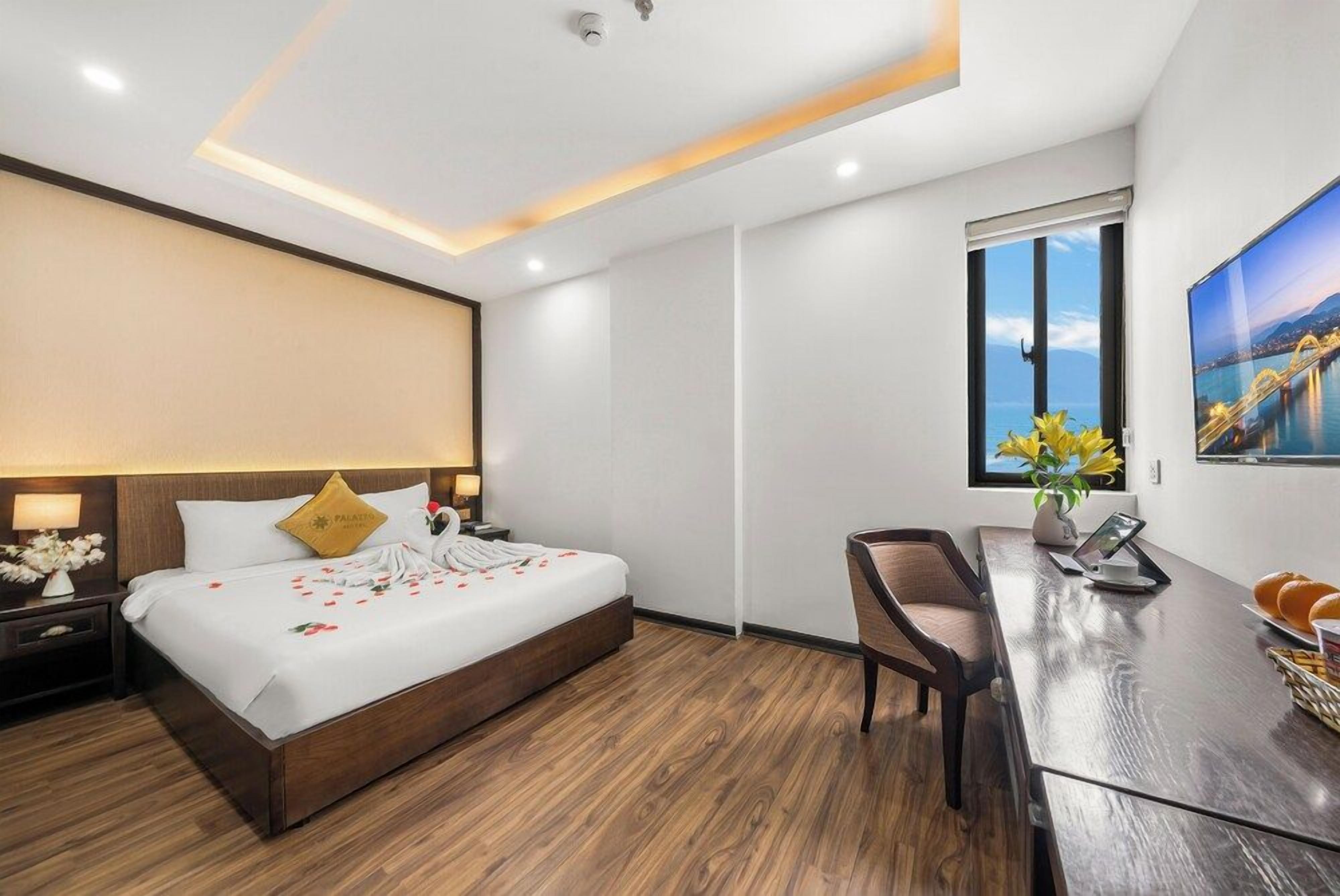 Quarto de hotel com cama, mesa de cabeceira, cadeira, escrivaninha e janela com vista para a cidade.