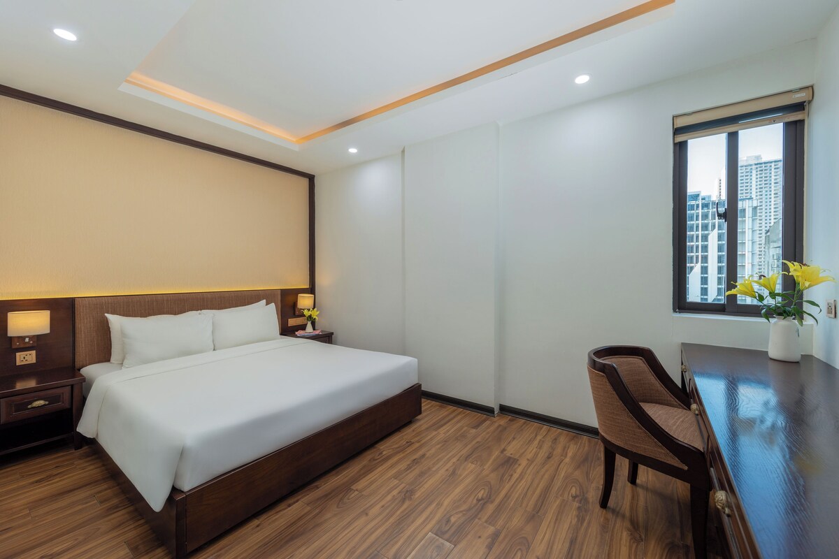 A hotel room with a large bed, wooden flooring, a desk with a chair, a window with a view of buildings, and a nightstand with a lamp.