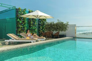 Uma área à beira da piscina com espreguiçadeiras, um guarda-sol e vista para a paisagem urbana.