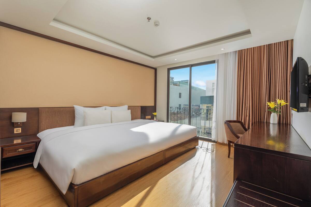 A hotel room with a large bed, wooden headboard, a desk with a flat-screen TV, and a balcony with a view of the city.