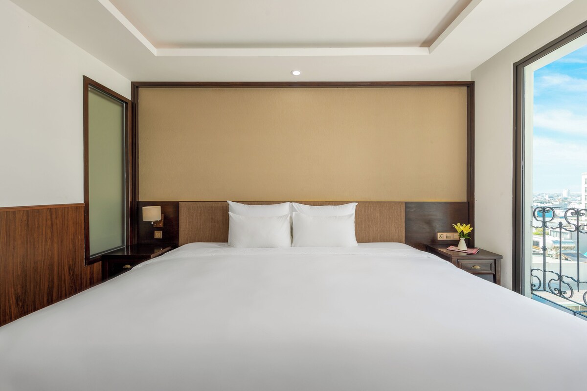 A hotel room with a large bed, wooden headboard, bedside tables, and a view of the city through a large window.