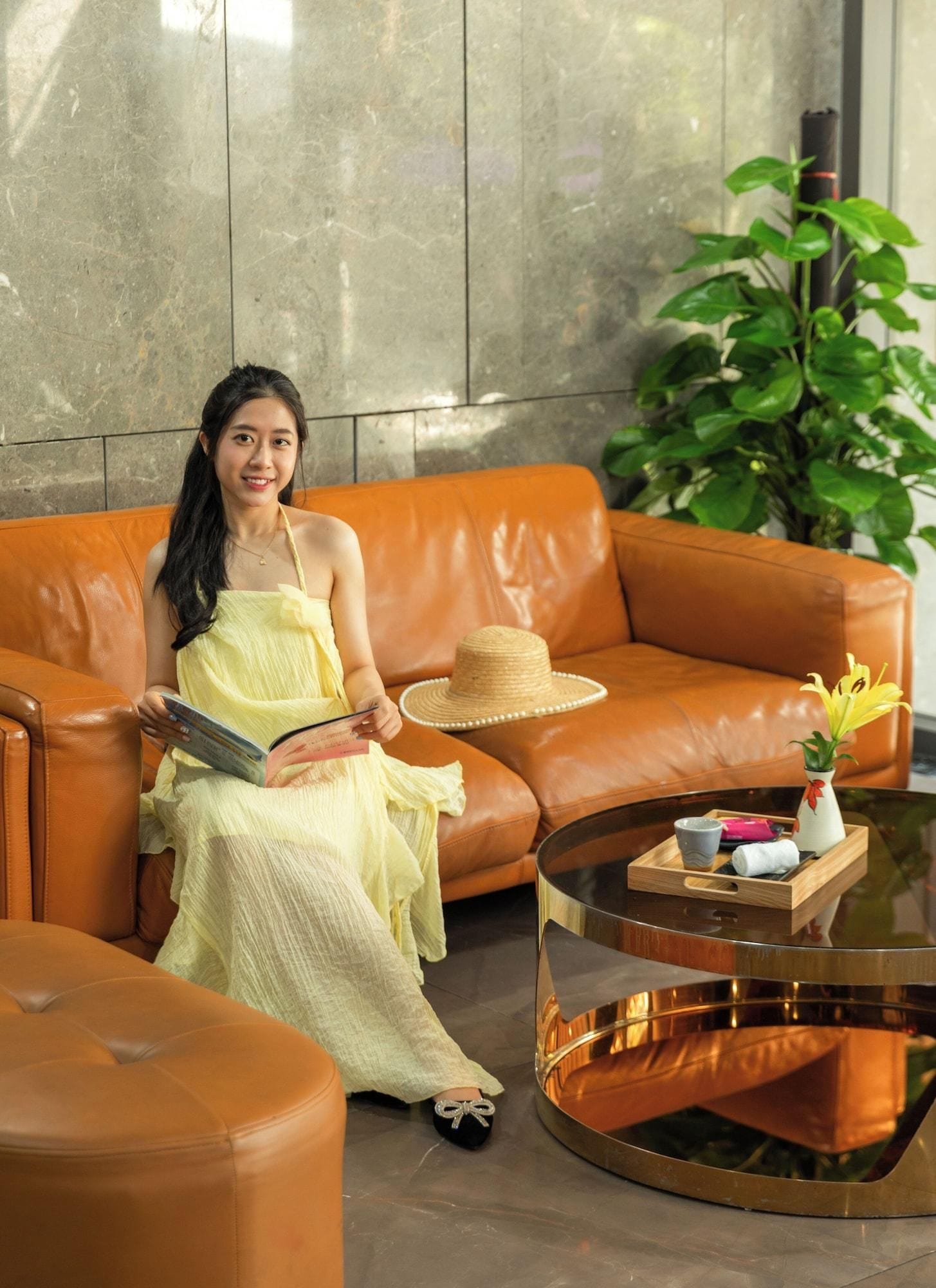 A woman sitting on a brown leather sofa in a modern room with a glass coffee table, a potted plant, and a small vase with flowers.