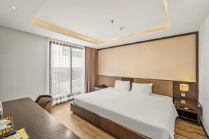A hotel room with a large bed, a desk, a chair, a bedside table, and a window with curtains. - Palazzo 3 (Da Nang Municipality)
