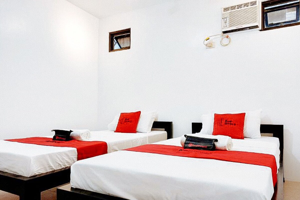 Une chambre d’hôtel avec deux lits, chacun agrémenté de linge de lit blanc et de touches de couleur rouge. On y trouve une petite fenêtre et un climatiseur fixé au mur.