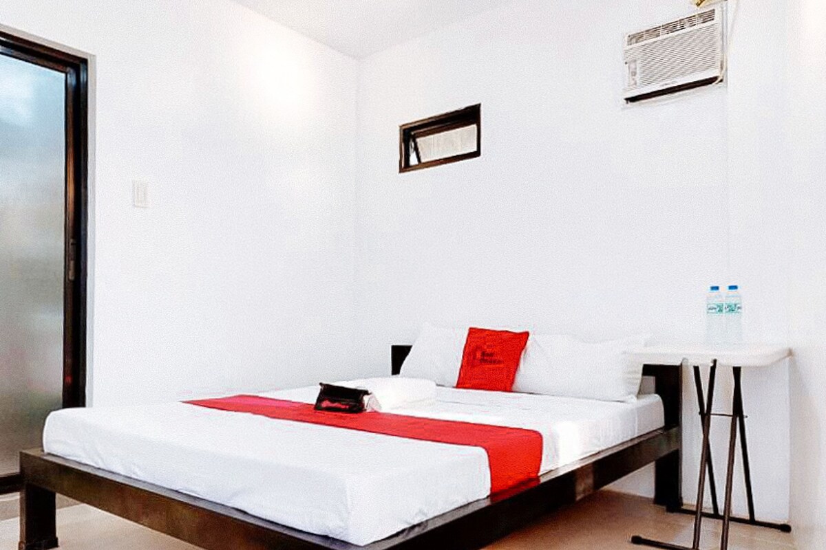Un lit simple avec une couvre-lit blanche et rouge, une petite table de chevet et une bouteille d’eau fixée au mur.