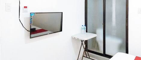 Habitación de hotel con cama, una mesita con silla, espejo, puerta corrediza de vidrio y toma de corriente.