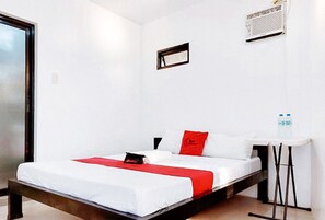 Un lit simple avec une couvre-lit blanche et rouge, une petite table de chevet et une bouteille d’eau fixée au mur.