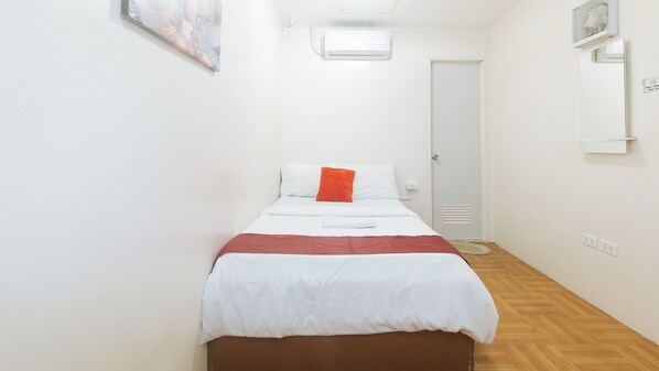 A small, narrow hotel room with a single bed, a red throw pillow, and a white headboard. There is a white air conditioning unit on the wall and a door with a mirror above it. - Check Inn Hotel Dumaguete City By Reddoorz (Dumaguete)