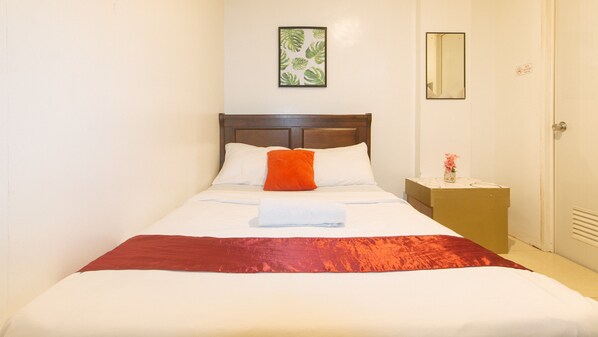 A neatly made bed with white and red bedding, a wooden headboard, a small bedside table with a vase of flowers, and a framed picture on the wall. - Check Inn Hotel Dumaguete City By Reddoorz (Dumaguete)