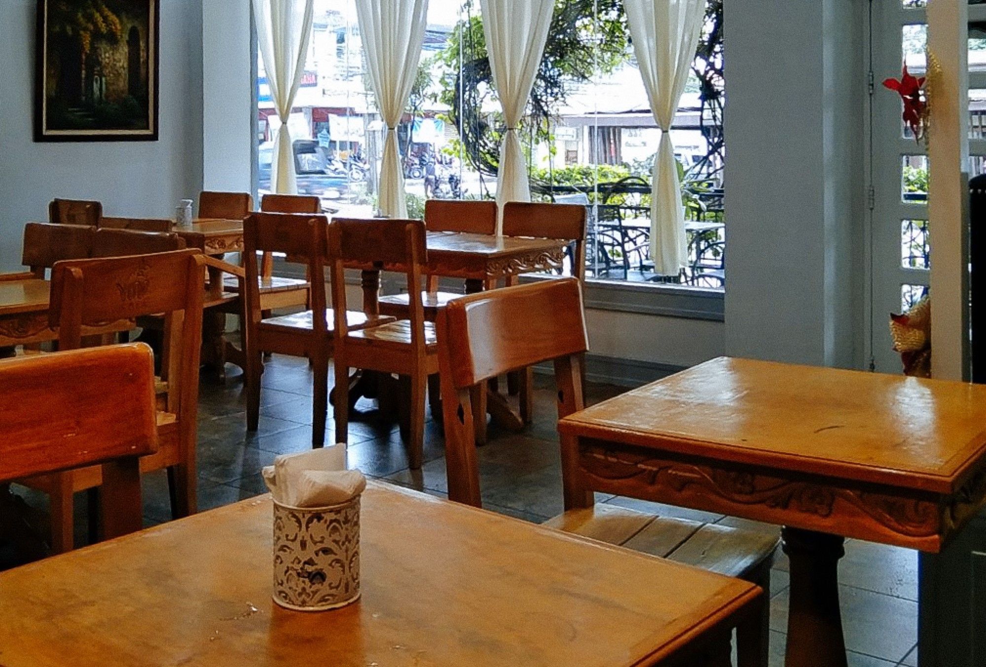 A restaurant interior with wooden tables and chairs, a painting on the wall, and a view of the street outside.